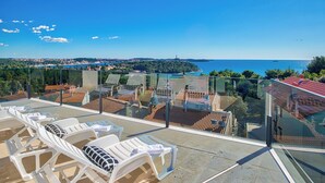 Eine Dachterrasse mit weißen Lounge-Sesseln und einer Glasbrüstung mit Blick auf eine Küstenstadt.