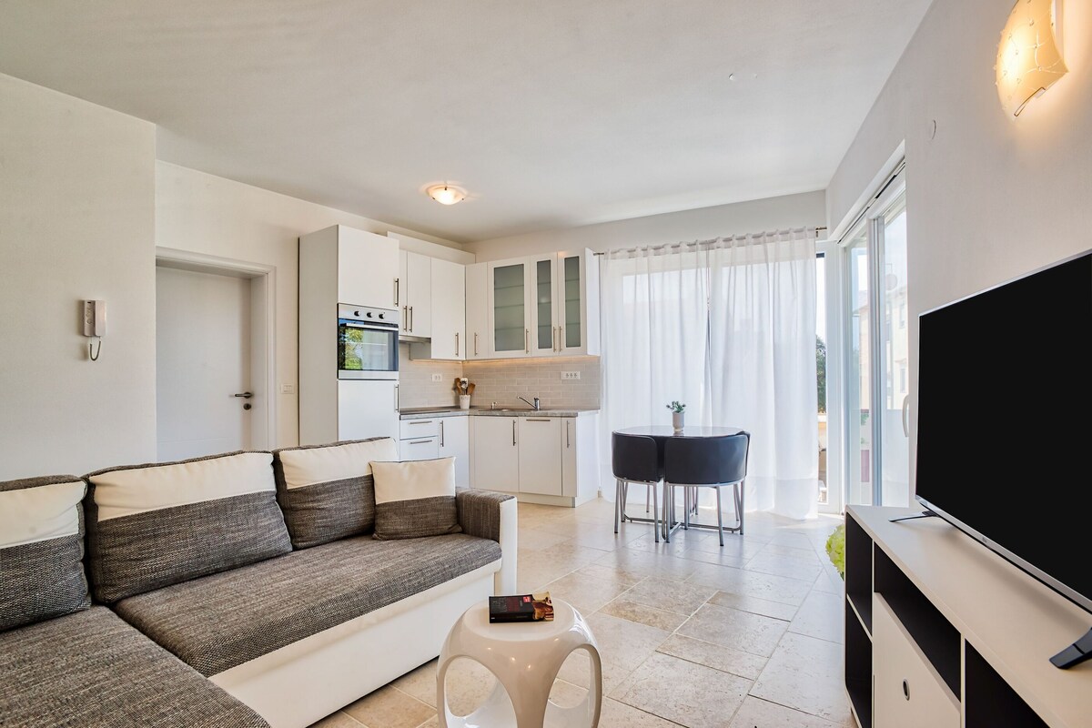 A modern living room with a sofa, a small white table, a TV, and a kitchen area with a dining table.