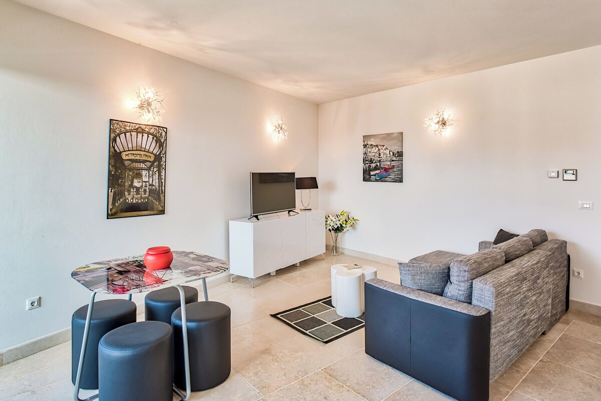 A modern living room with a sofa, a dining table with stools, a TV on a white cabinet, and artwork on the walls.