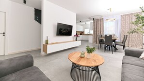 A modern living room with a flat-screen TV, a wooden coffee table, grey sofas, and a dining area with a table and chairs. - Villa Flora (Kastela)