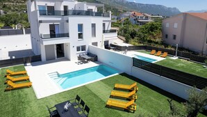 A modern two-story building with a swimming pool, outdoor seating, and a view of mountains. - Villa Flora (Kastela)