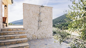 A stone-walled outdoor shower area with steps leading up to it, surrounded by natural vegetation and a mountainous backdrop. - Villa Maslina (Marina)