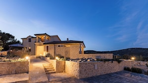 A modern house with a stone facade, a well-lit entrance, and a paved driveway. - Villa Maslina (Marina)