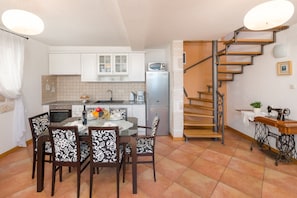 Cocina compacta con armarios blancos, un refrigerador y un comedor con sillas estampadas. Una escalera en espiral conduce a un nivel superior.