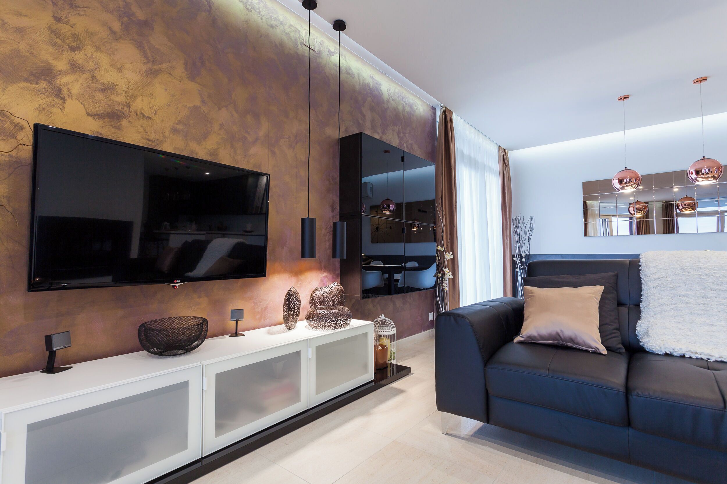 A modern living room with a flat-screen TV mounted on the wall, a dark-colored sofa with cushions, a white console table with decorative items, and a large mirror reflecting the room.