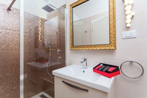 Une salle de bain avec une douche à parois de verre, un lavabo blanc avec un miroir à cadre doré et une boîte à produits de toilette rouge posée sur le lavabo.