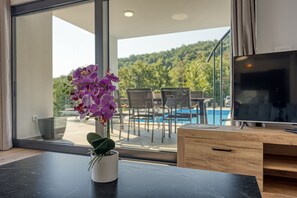 A modern room with a TV, a wooden desk, a potted orchid, and a view of a pool and greenery outside. - Villa Nono (Karojba)