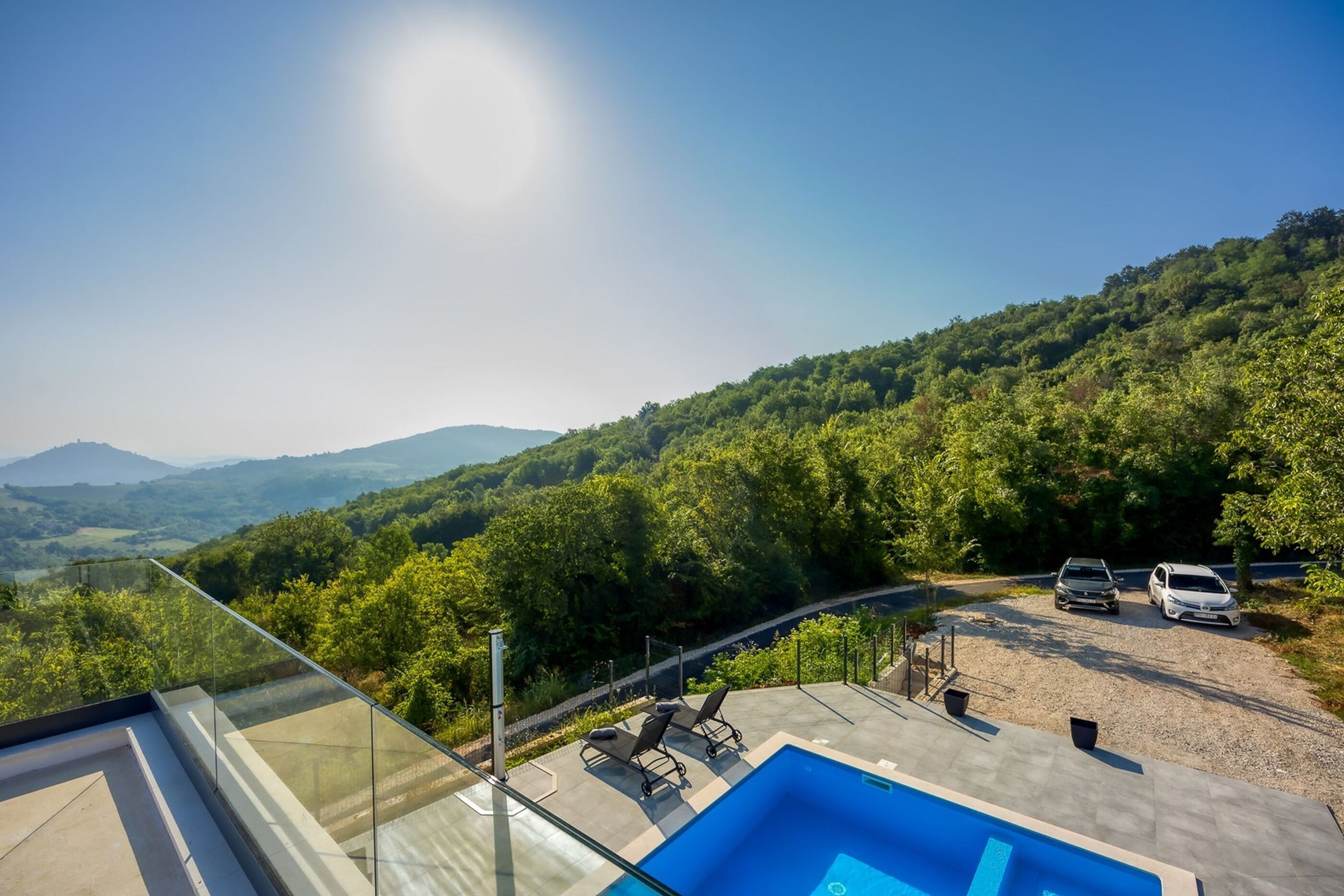 A pool with a glass balustrade, a carport with two cars, and a view of a forested hillside.