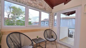 Ein Balkon mit zwei schwarzen Korbstühlen, einem kleinen Tisch mit einer Pflanze und Blick auf Gebäude durch große Fenster.