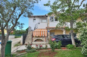 A two-story building with a stone facade, a small outdoor seating area with a striped awning, and a parked car in front. - Apartment Emilija (Fazana)