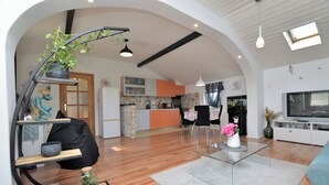 Ein modernes Wohnzimmer mit einem Glastisch, einer Couch und einem Fernseher. Im Hintergrund ist eine Küche mit orangefarbenen Schränken und einem weißen Kühlschrank zu sehen.