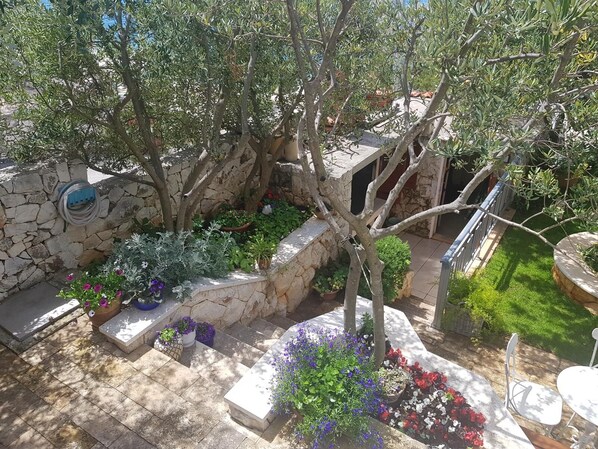A stone-walled courtyard with a variety of potted plants and flowers, a stone bench, and a small table. - Apartments Roza (Rogoznica)