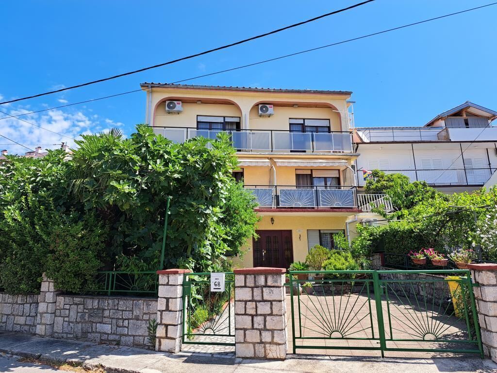 A two-story residential building with a balcony on the upper floor, a stone wall in front, and a green gate.