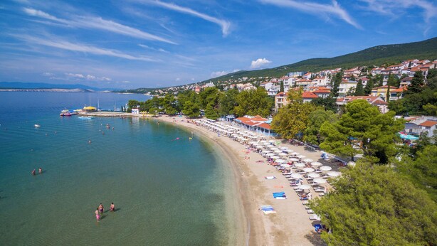 Egy tengerparti város, ahol a strandot napernyők és strandhúzók szegélyezik, a tenger kristálytiszta és kék, az épületek pedig piros tetővel rendelkeznek.