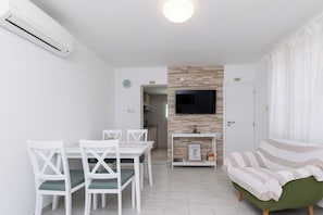 A modern living room with a green sofa, a white dining table with chairs, a flat-screen TV mounted on a wall, and a small shelf with decorative items. - House Zeljko (Rab)