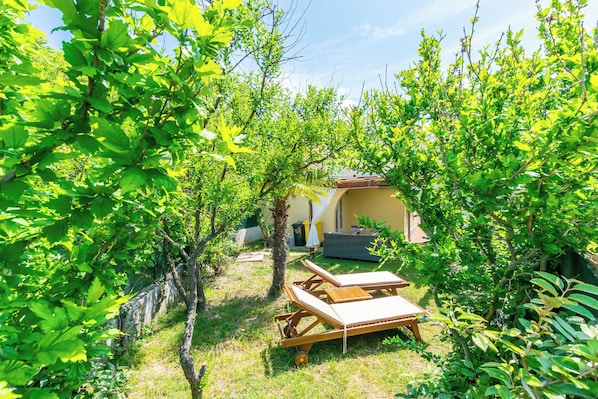A garden with a sun lounger and a table, surrounded by lush greenery and a building in the background. - House Zeljko (Rab)