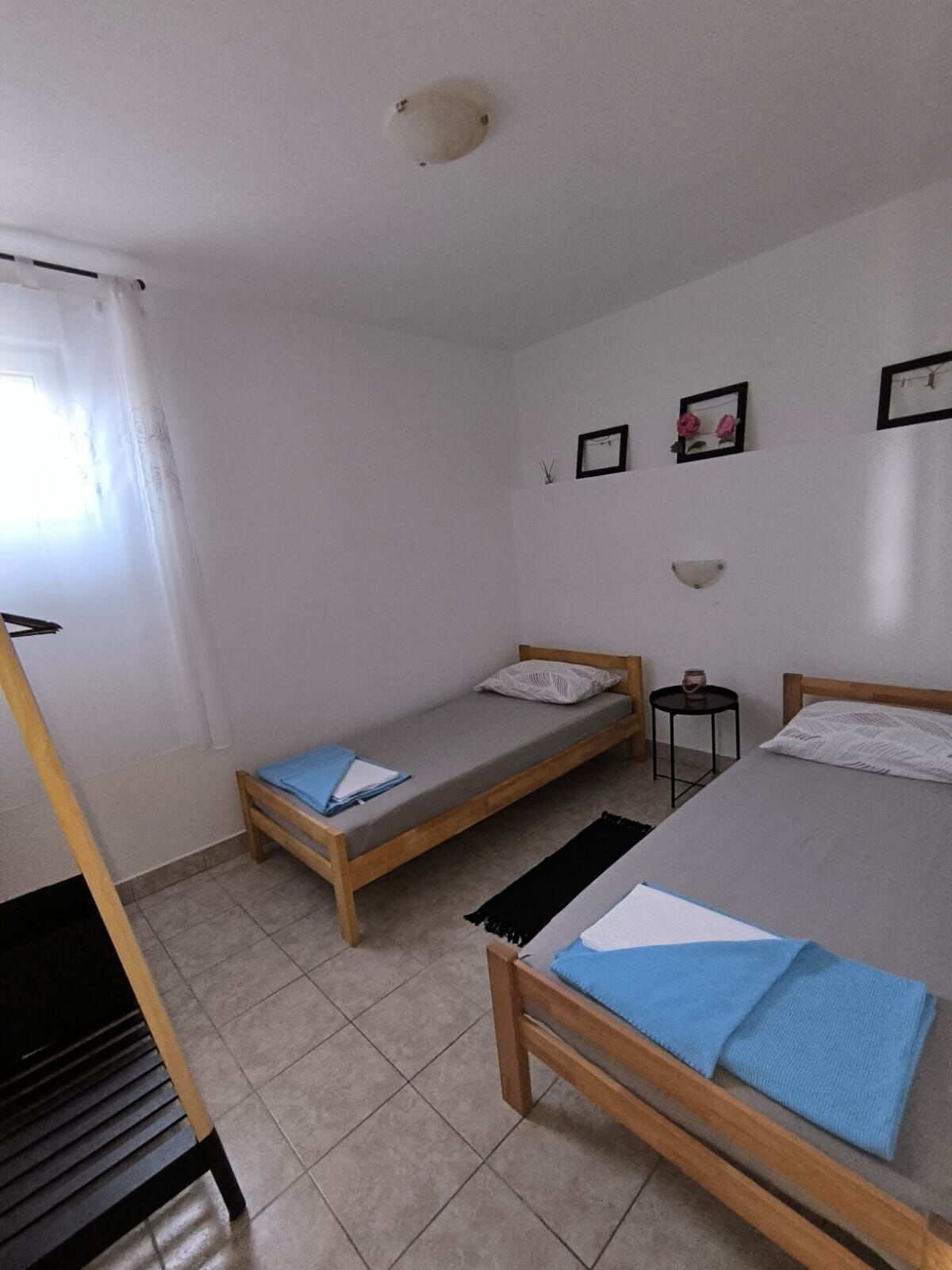 A small, clean room with two single beds, a wooden bench, a small table, and a window with curtains.