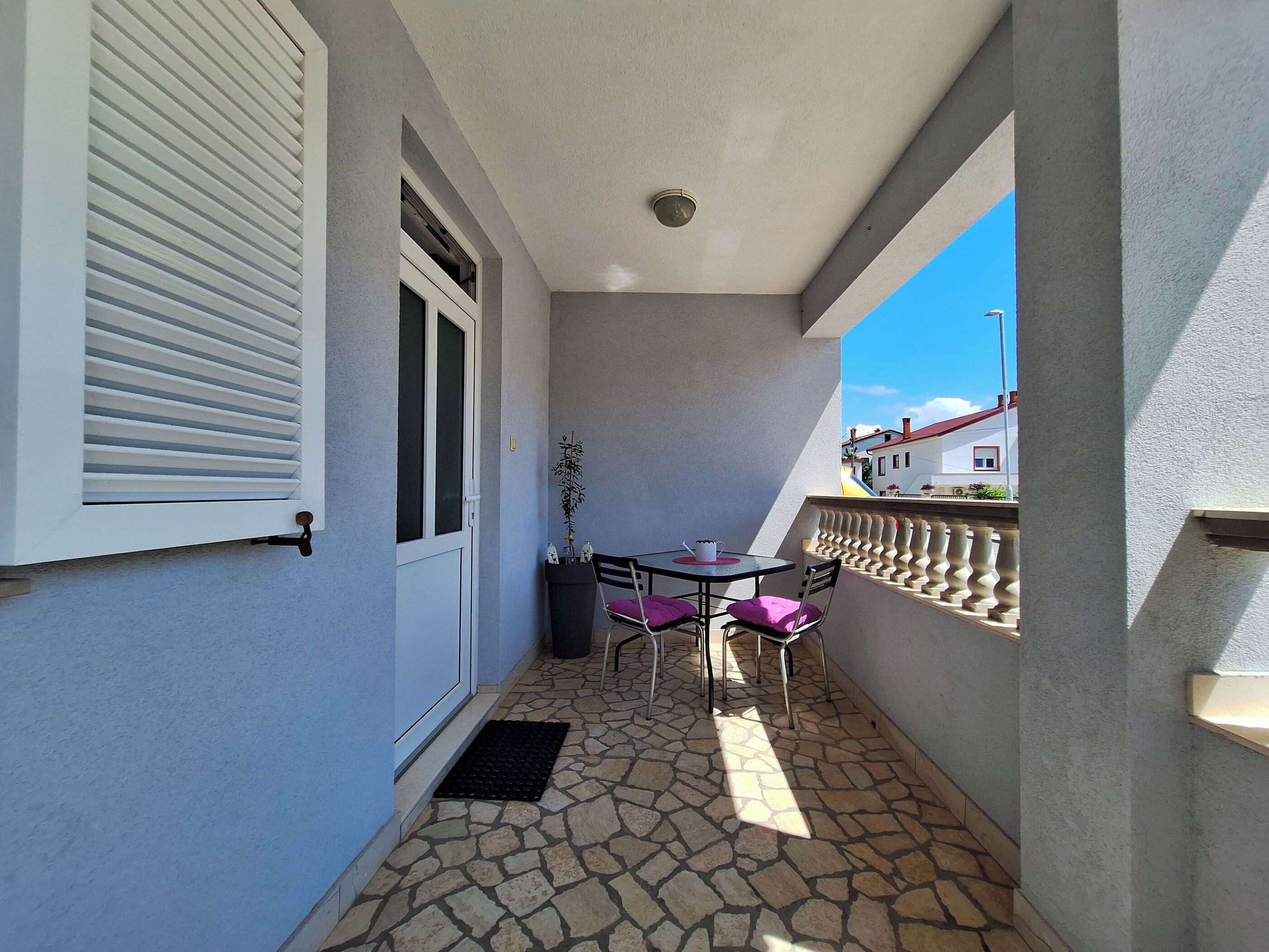 A small balcony with a table and chairs, a potted plant, and a view of the sky and buildings.