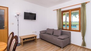 Ένα σαλόνι με έναν καναπέ, μια τηλεόραση, ένα φωτιστικό, ένα τραπέζι φαγητού και ένα παράθυρο με κουρτίνες.