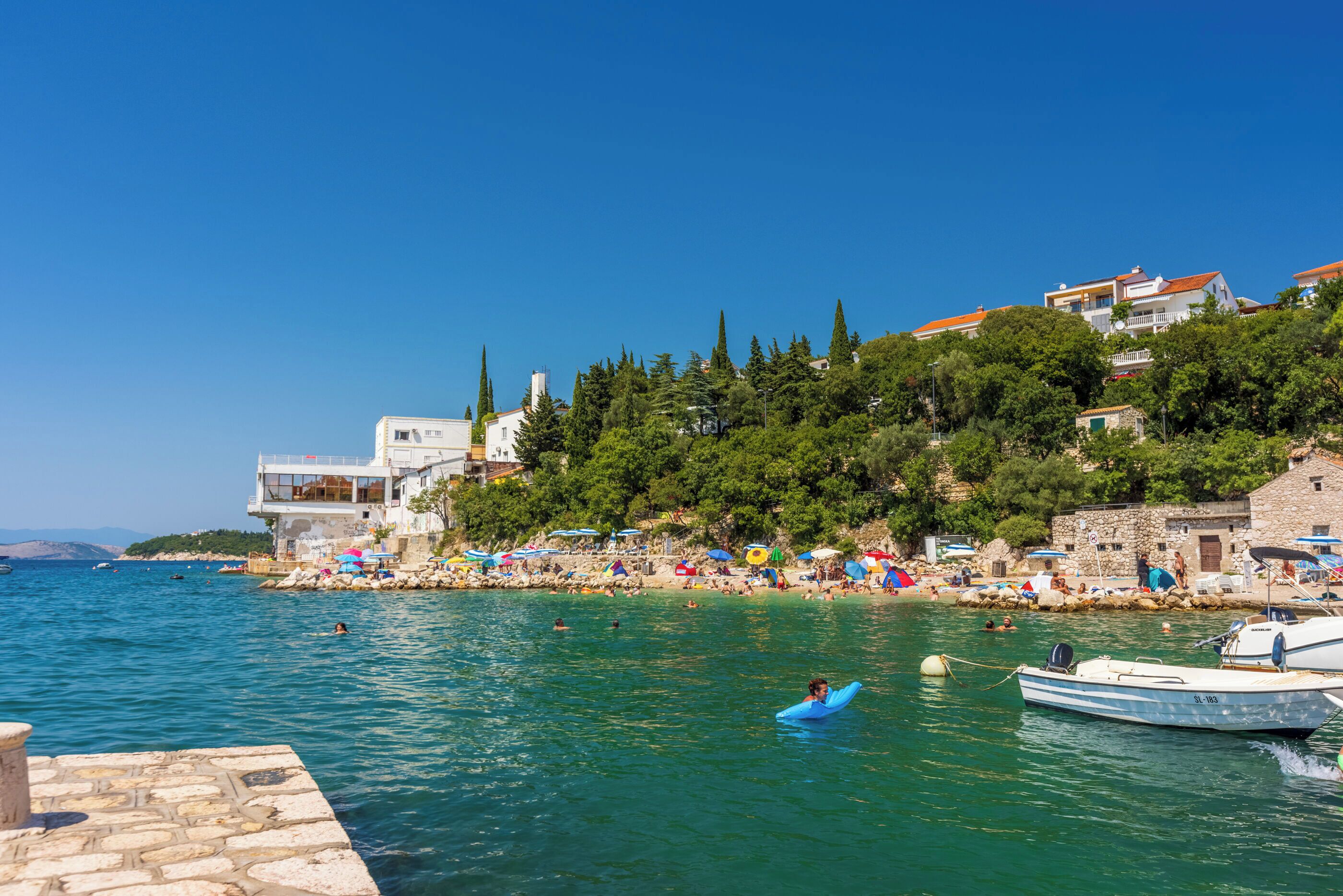A coastal town with a beach, clear blue sky, and a person on a blue surfboard.