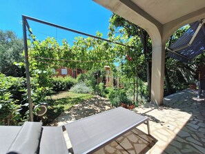 Un patio abrité, doté d’une table et de chaises, entouré d’une végétation luxuriante et surplombé d’un ciel bleu azur.