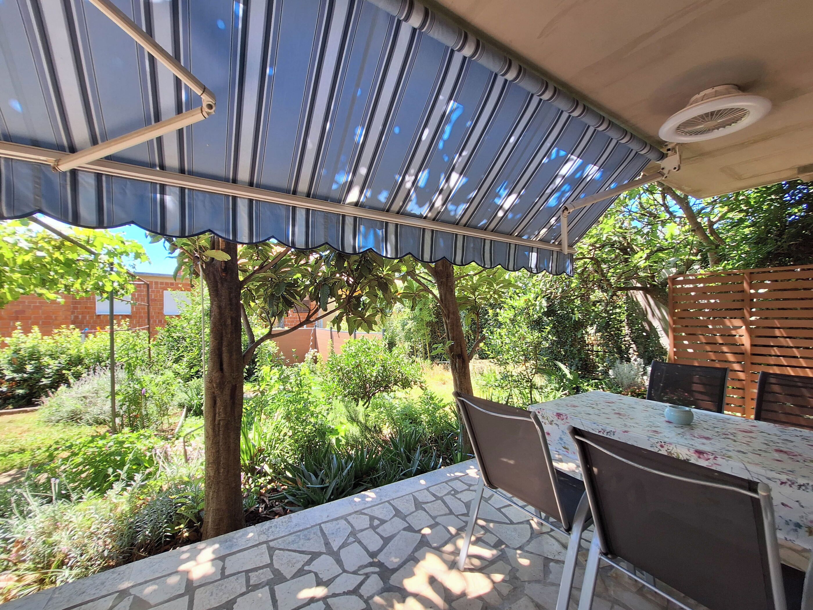 Un patio exterior con una mesa y sillas cubiertas, rodeado de exuberante vegetación y un muro de ladrillo.
