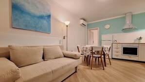 Ett vardagsrum med en beige soffa, ett matbord med stolar, ett kök med vita vitvaror och en blå målning på väggen.