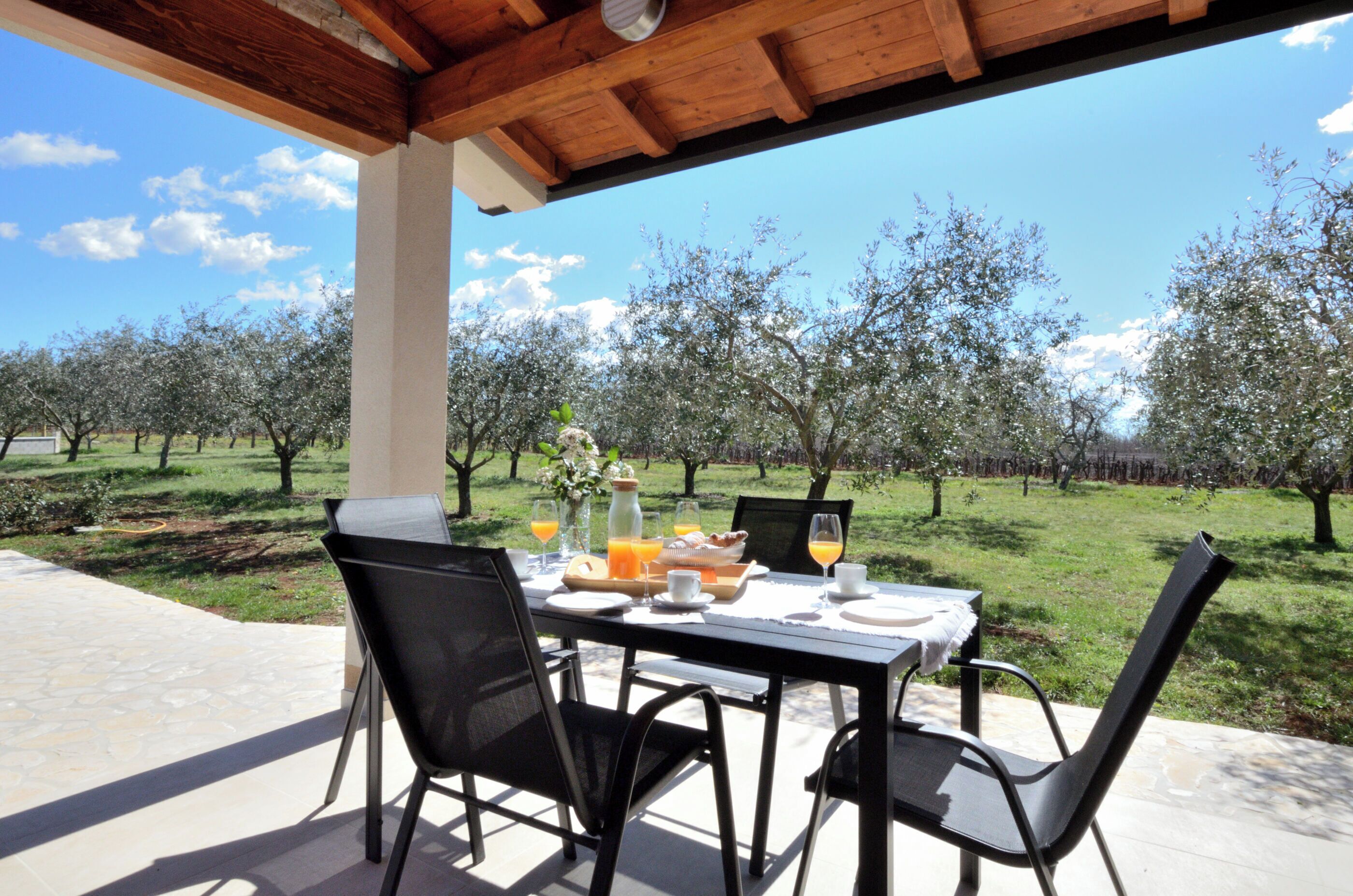 An outdoor dining area with a table set for two, chairs, and a view of an orchard.