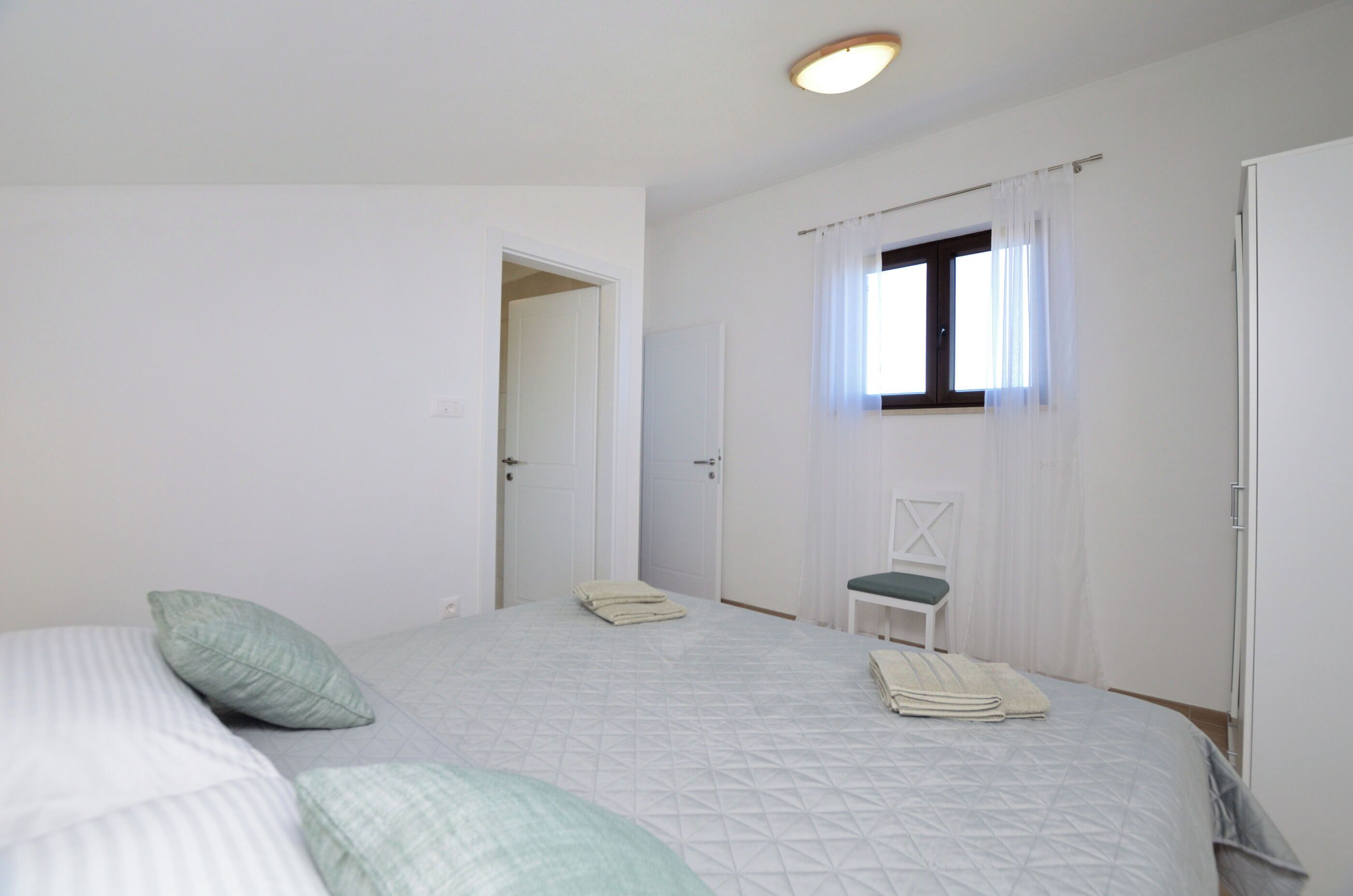 A bedroom with a bed, a chair, a window with curtains, and a door.