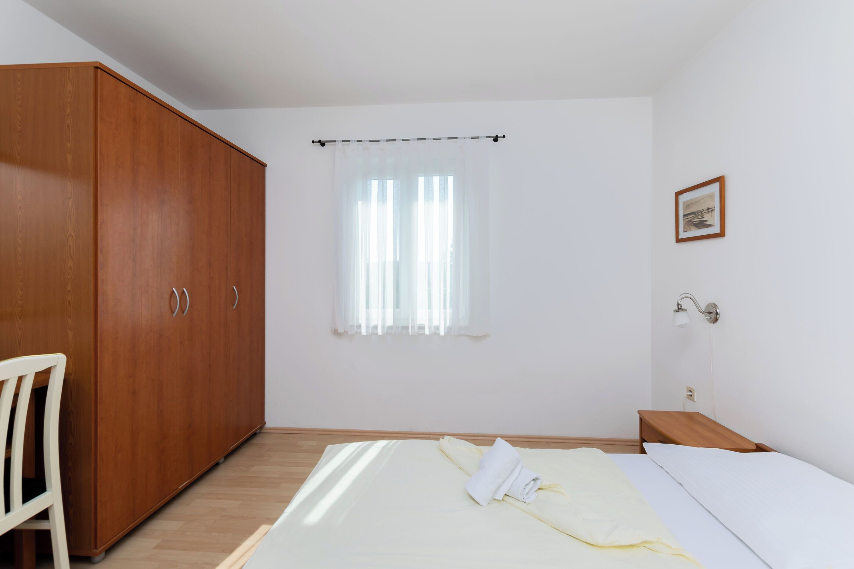 Een slaapkamer met een bed, een houten garderobe, een stoel, een nachtkastje, een lamp en een raam met gordijnen.