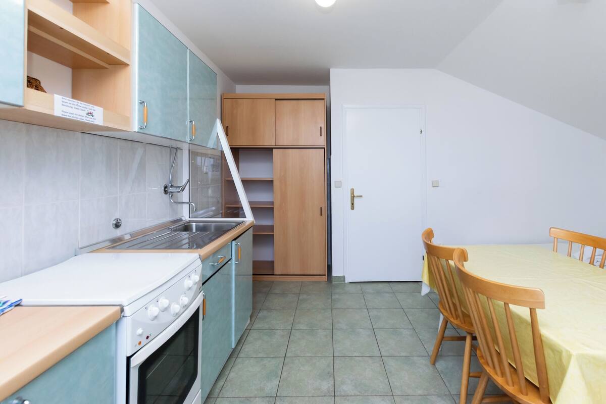Une cuisine compacte équipée d’une cuisinière, d’un évier et de meubles en bois. On y trouve une table à manger avec des chaises, et une porte blanche en arrière-plan.