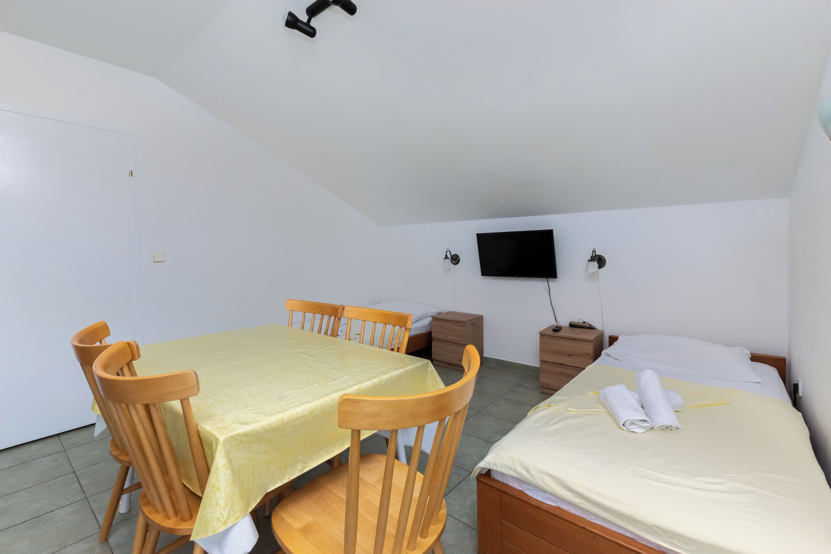 Una habitación con una mesa de comedor, sillas, una cama con colchón y almohadas blancas, un televisor en la pared y un ventilador de techo.