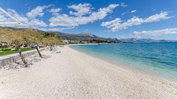 Ein Strand mit klarem, blauem Wasser, Kiesufer und Bergen im Hintergrund.