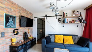 Een woonkamer met een blauwe bank, een houten salontafel, een aan een bakstenen muur bevestigde televisie, en een boekenkast met boeken en decoratieve objecten.