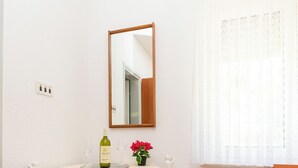 A small, neatly arranged room with a round table, a chair, a wooden nightstand, a bed, a mirror, and a window with sheer curtains. - Apartment Jele (Omis)