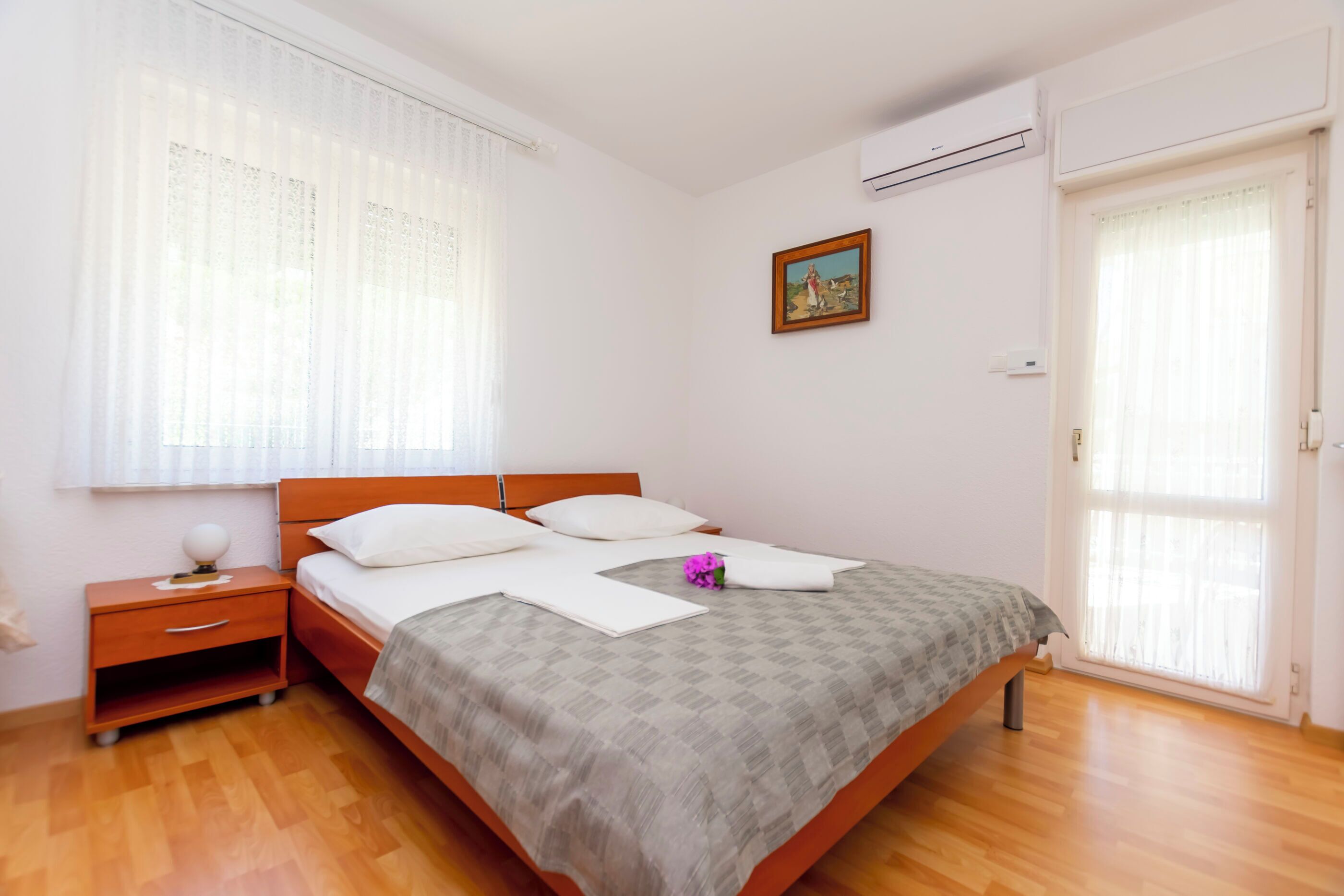 A bedroom with a wooden bed, white curtains, a small bedside table, and a single wall-mounted air conditioner.
