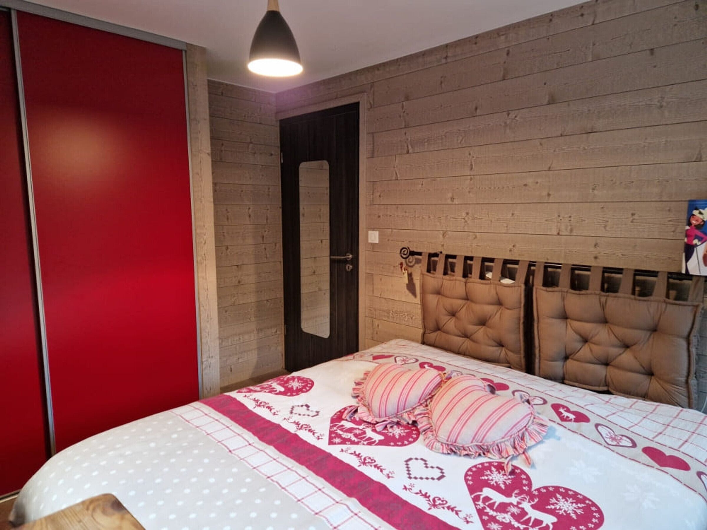 A bedroom with a wooden headboard, a bed with a heart-patterned bedspread, a red wardrobe, and a door.