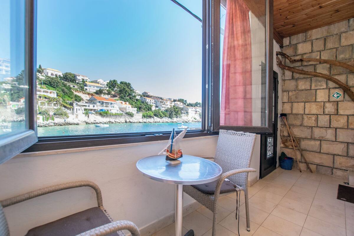 A balcony with a view of a coastal town, a small table with a model ship, wicker chairs, and a stone wall.