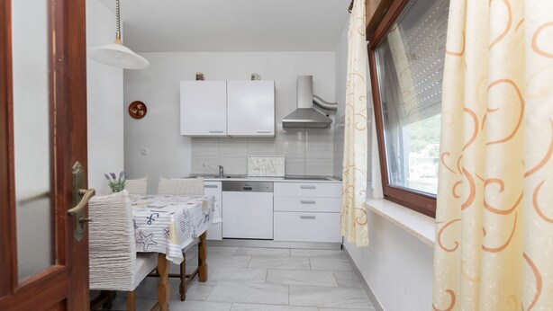 A compact kitchen with white cabinets, a built-in stove, and a window with patterned curtains. - Apartments First row (Korčula)