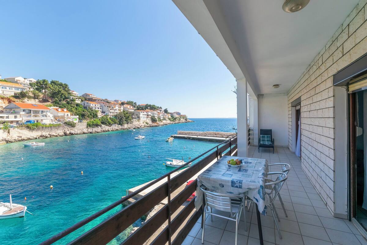 A balcony with a table and chairs overlooking a coastal area with boats and buildings.