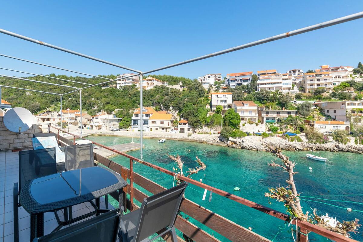 A balcony with a table and chairs overlooking a coastal town with clear turquoise waters and buildings on a hillside.