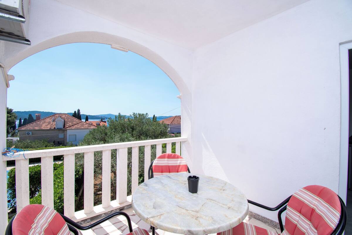 A balcony with a round table and chairs, a white railing, and a view of houses and trees.