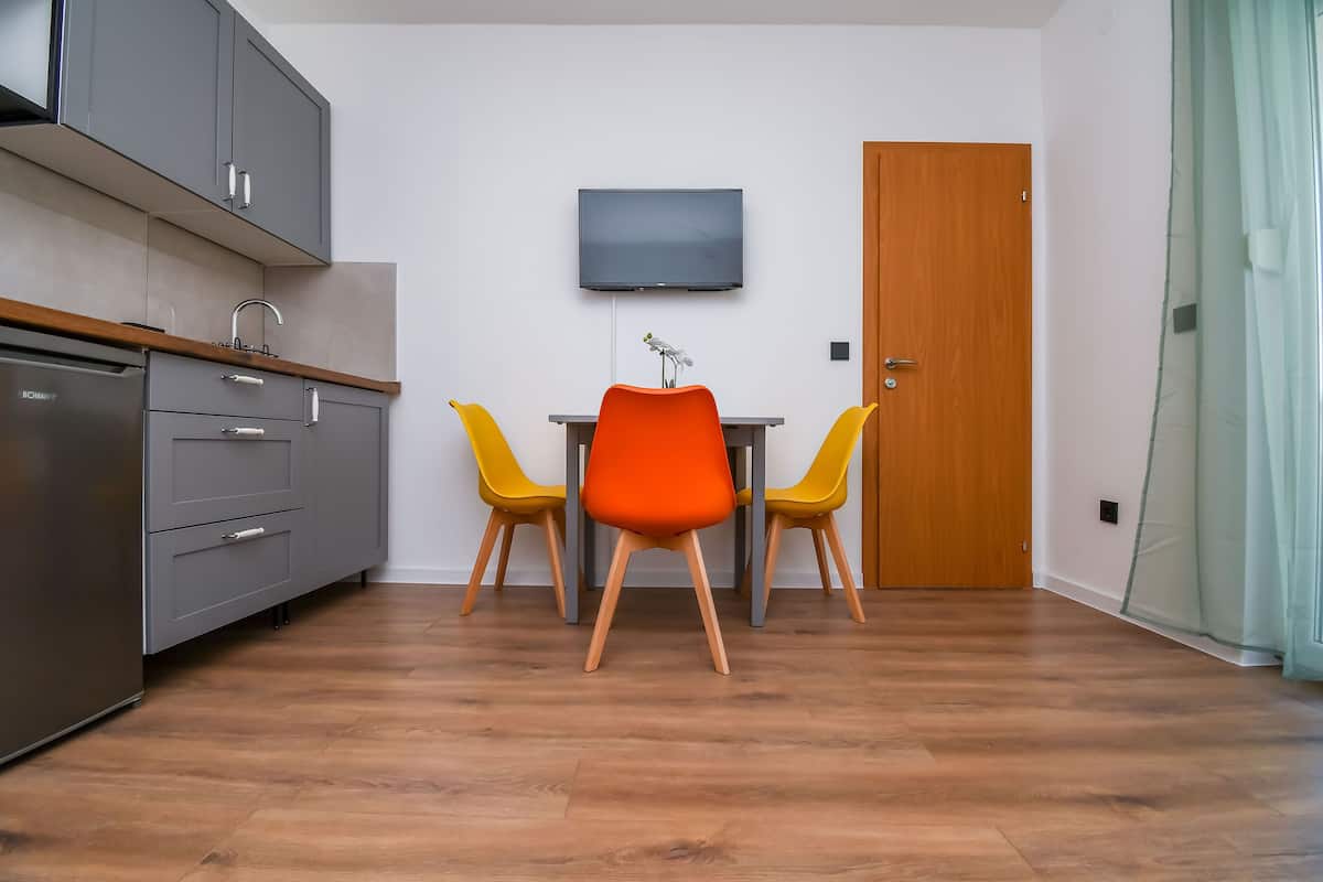 A compact kitchen with grey cabinets, a built-in oven, and a wooden countertop. There is a table with two yellow chairs and a single orange chair. A flat-screen TV is mounted on the wall above the table.