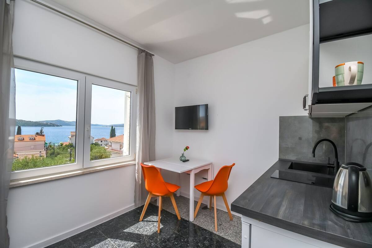 A compact kitchen with a white table and two orange chairs, a flat-screen TV mounted above it, and a view of the sea through large windows.