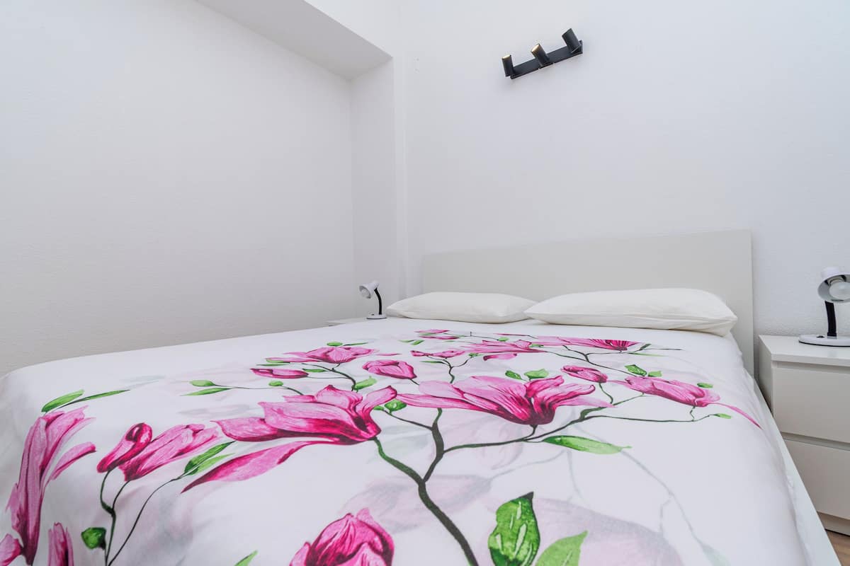 A bed with a floral bedspread, a white headboard, a bedside table with a lamp, and a wall-mounted light fixture.