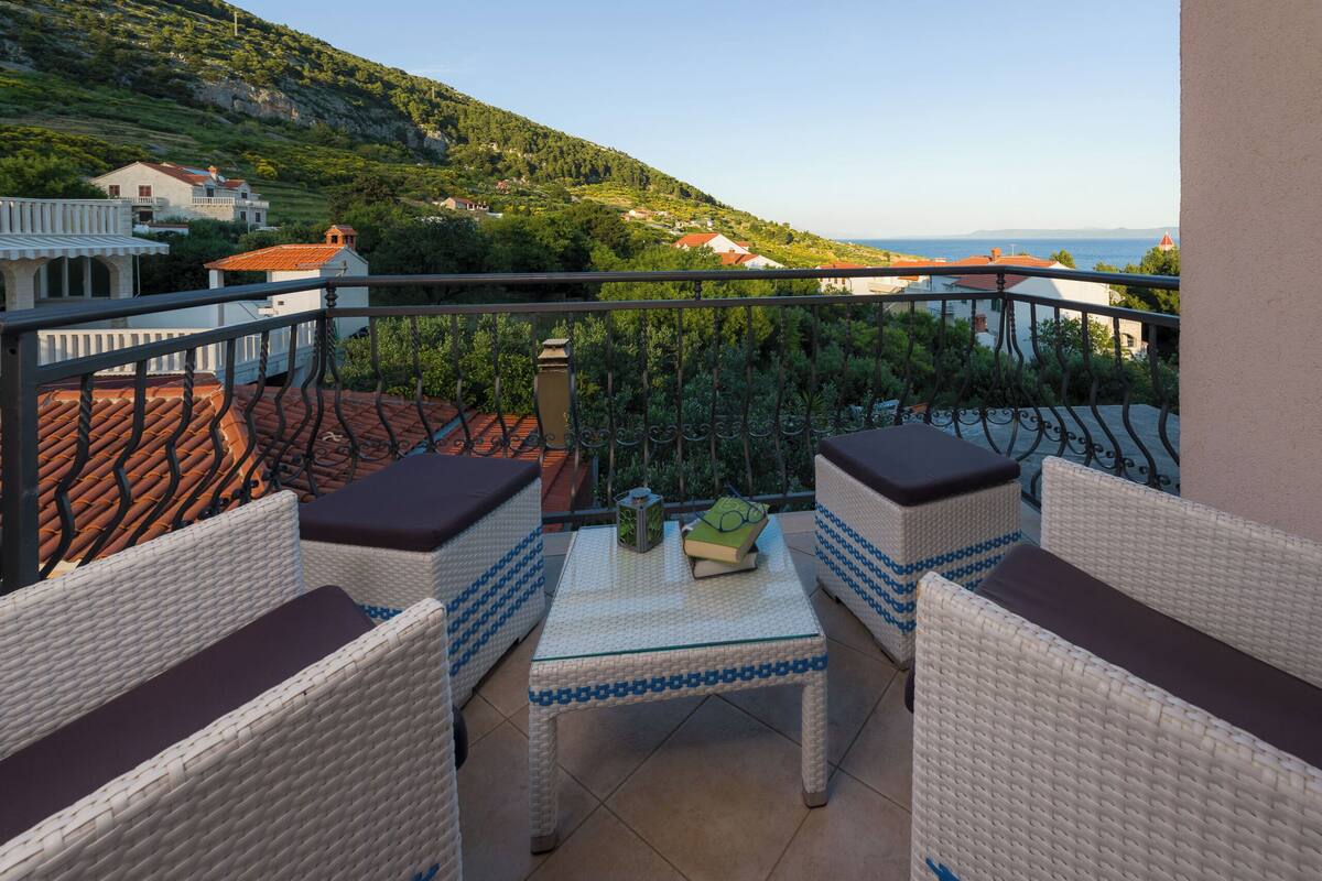 A balcony with wicker furniture, a glass table, and a view of a hillside with houses.