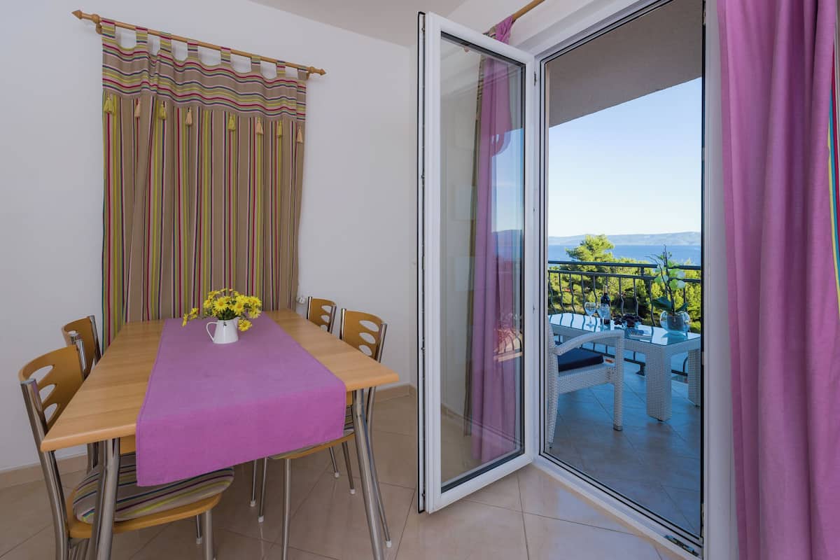 A dining area with a table set for six, a vase of yellow flowers, and a view of an outdoor balcony with wicker furniture.