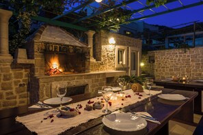 Un área de comedor al aire libre con chimenea de piedra, una mesa para dos con copas de vino y velas, y una pérgola sobre ella.