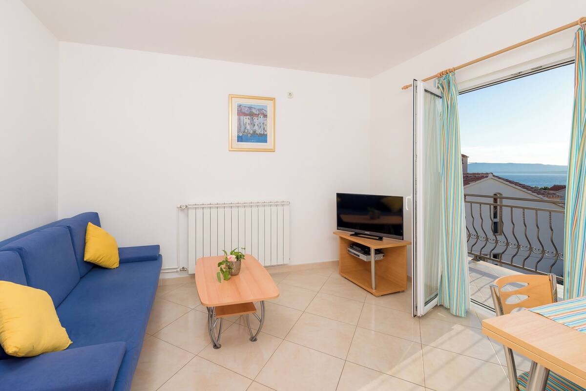 A modern living room with a blue sofa, a wooden coffee table with a plant, a TV on a stand, and a balcony with striped curtains.