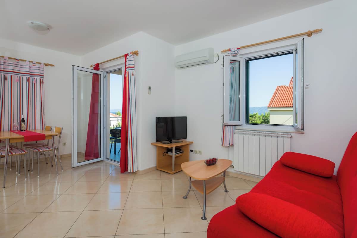 A room with a red sofa, a dining table with chairs, a TV on a stand, and a window with a view of buildings and hills.